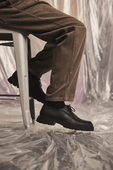 Schwarze Schuhe und beige Hosenbeine, sitzend auf einem Stuhl vor einem dekorativen Hintergrund.